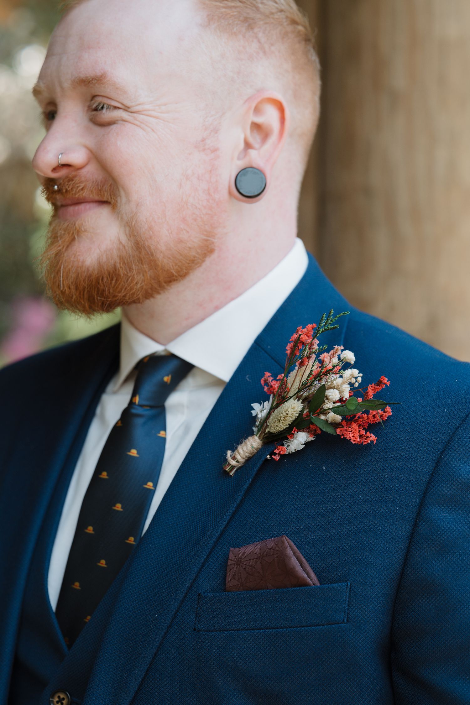 Red-haired groom in navy suit with floral boutonniere and gauge earring smiling on wedding day.