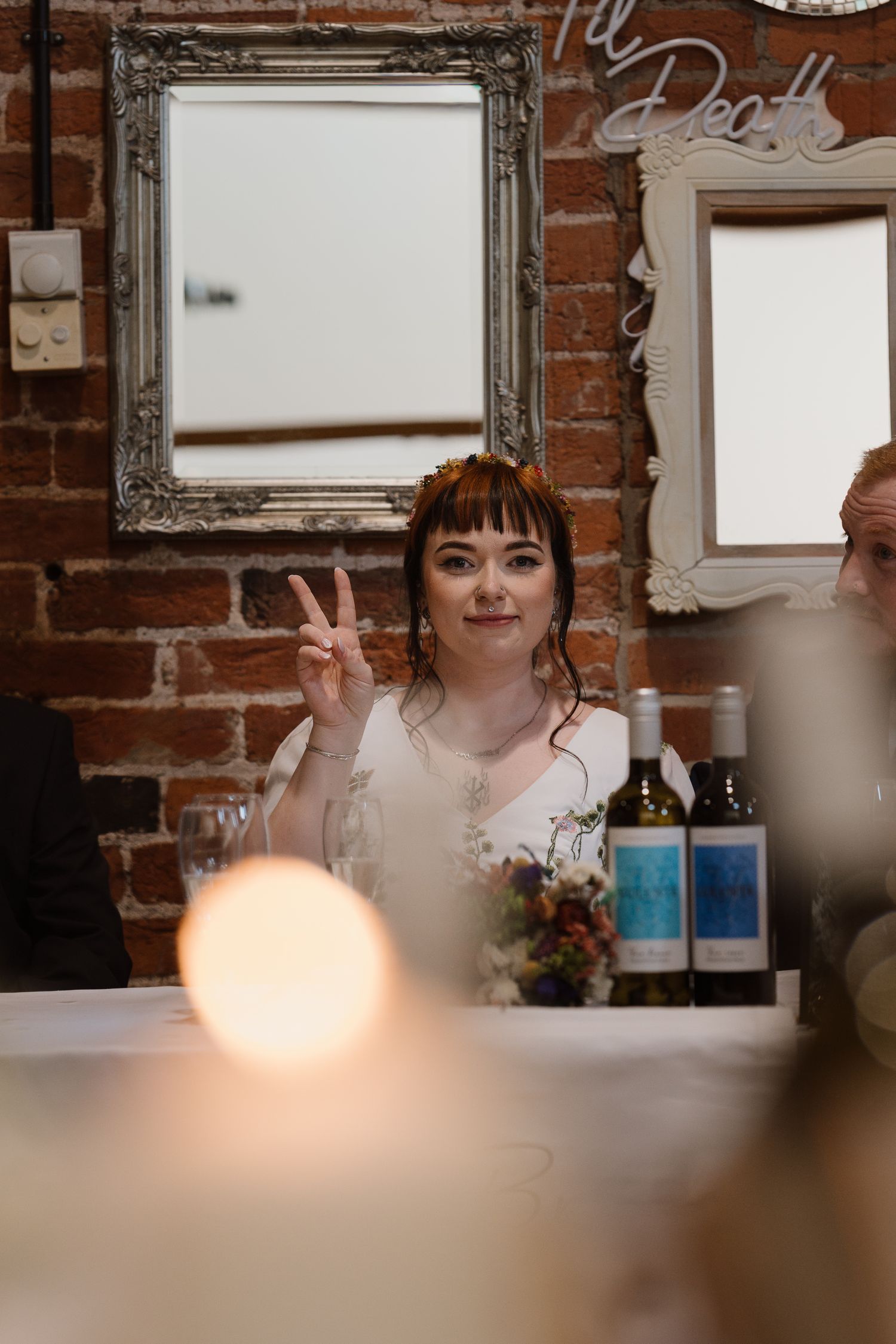Bride in white dress giving peace sign at wedding reception table with wine bottles and brick wall backdrop.