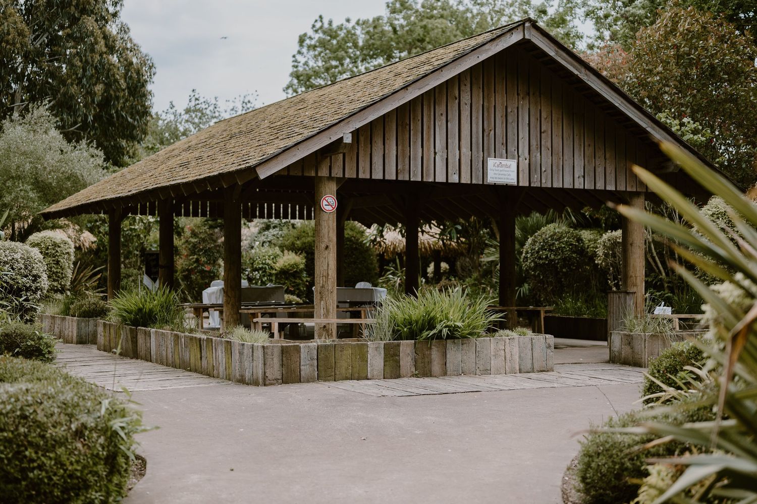 Rustic wooden open-air pavilion surrounded by lush greenery and manicured shrubs in a peaceful garden setting.