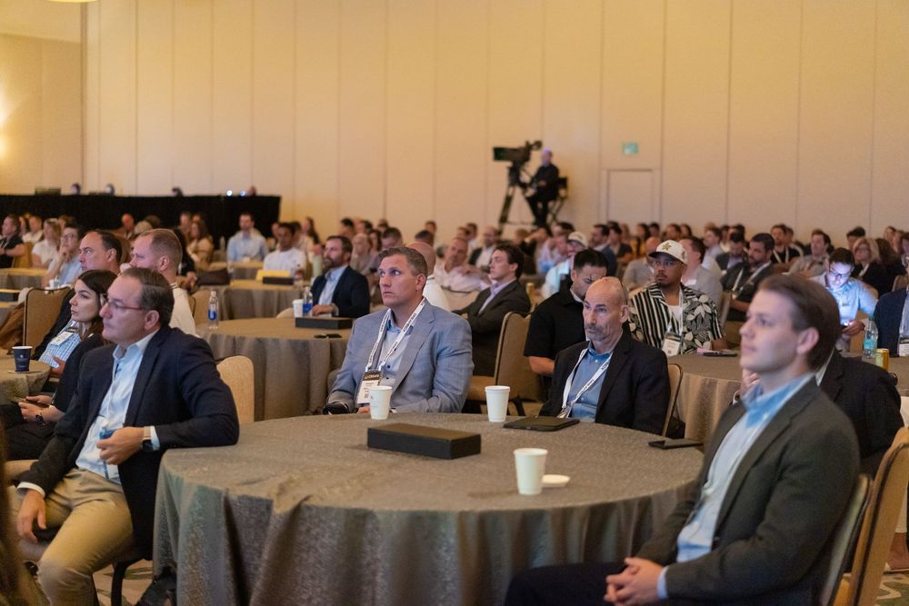 Business professionals seated at round tables attending a corporate conference in a large hotel ballroom.