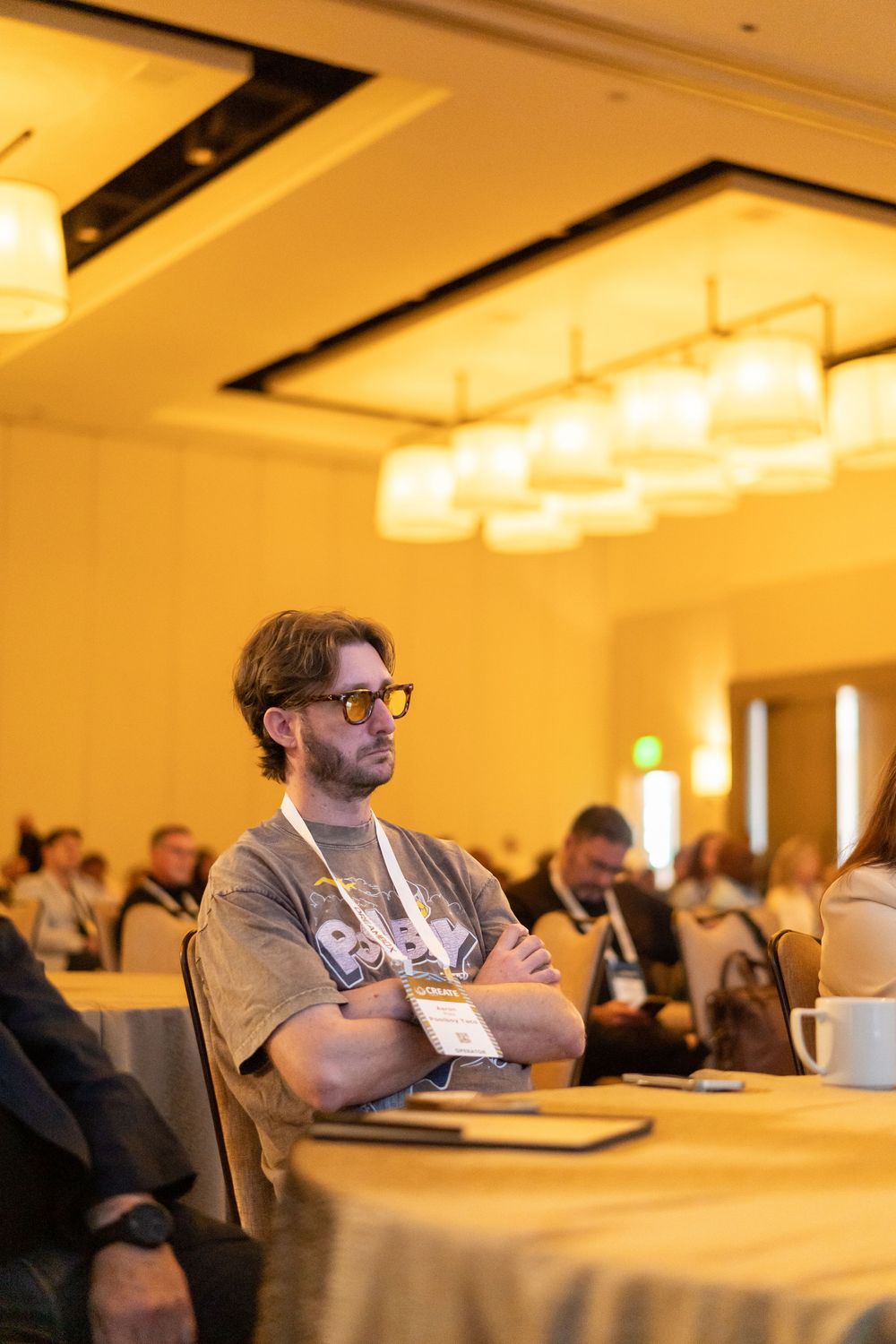 Man with yellow sunglasses and crossed arms sits attentively at a conference table in a warmly lit ballroom.