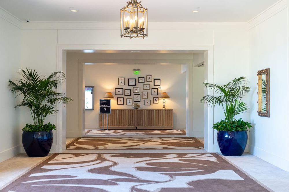 Elegant hotel lobby entrance with tropical plants, patterned area rug, framed wall art, and a decorative chandelier.