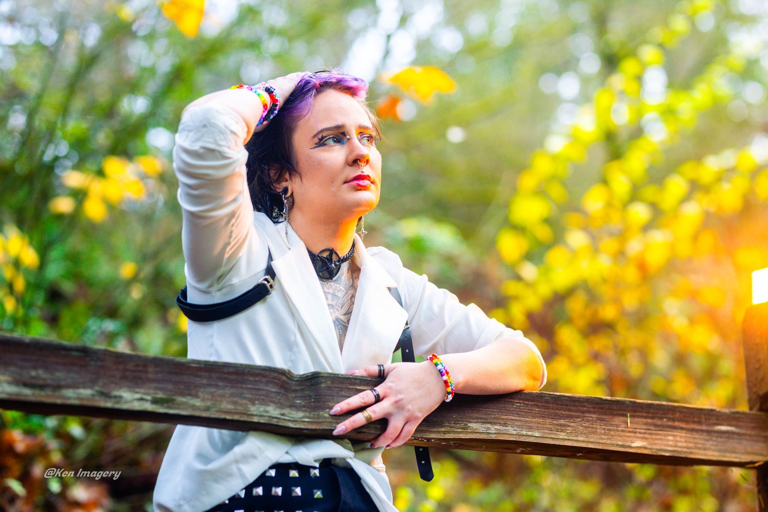 Woman with purple hair in white jacket leans on wooden fence surrounded by yellow autumn foliage outdoors.