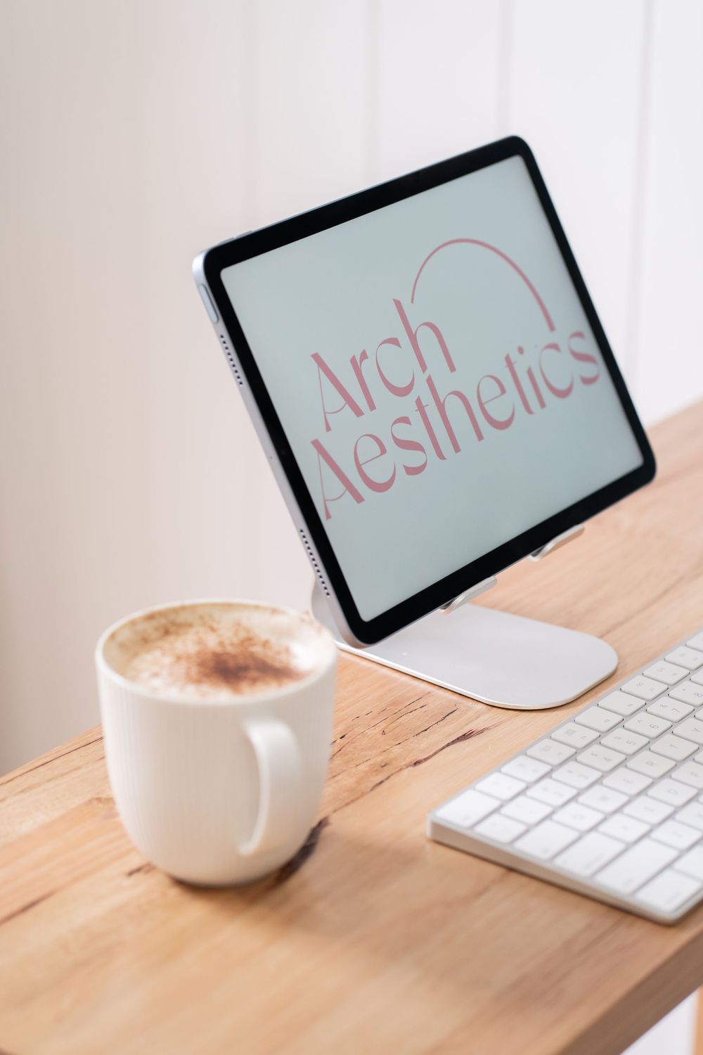 Tablet displaying Arch Aesthetics logo on a wooden desk with a cappuccino and white keyboard.