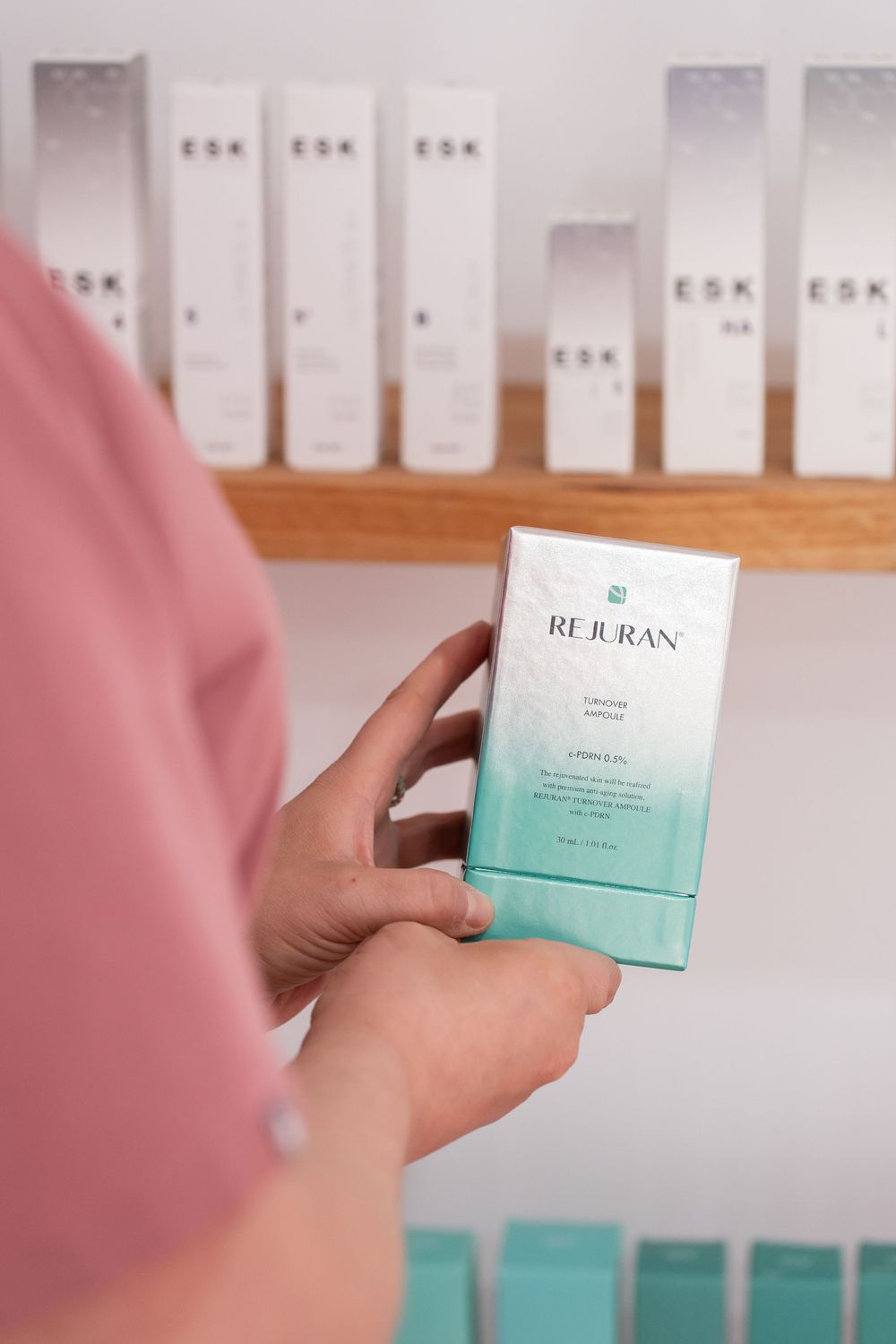 Person holding Rejuran skincare product box in front of ESK products on wooden shelf.