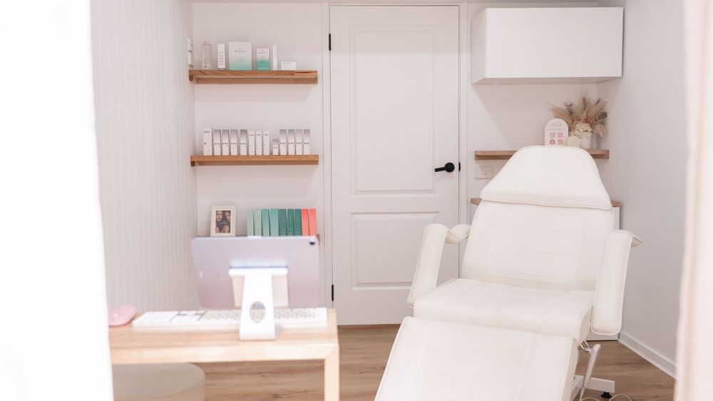 Bright white spa treatment room with reclining chair, product shelves, and minimalist decor.