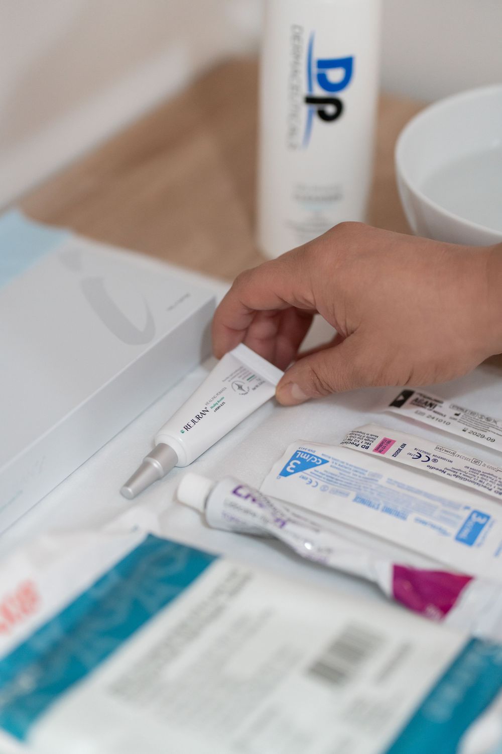 Hand holding a small medical cream tube near skincare products and bandages on a white surface.