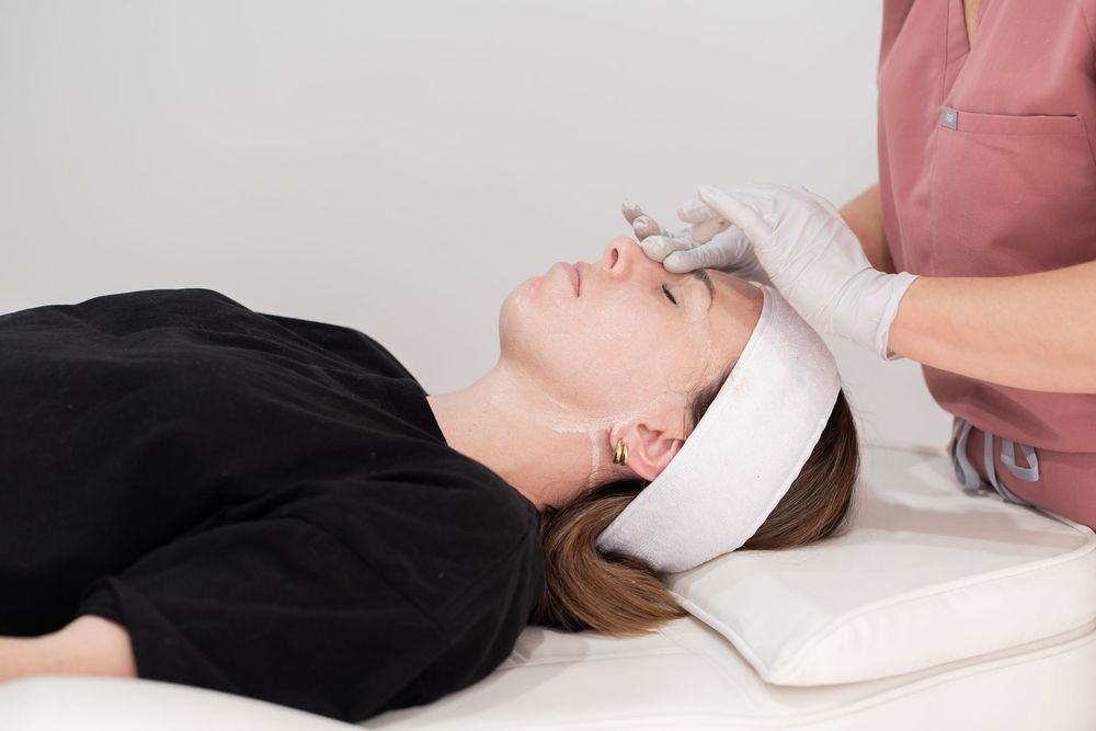 Esthetician in pink scrubs and gloves performing a facial treatment on a woman lying on a spa bed.