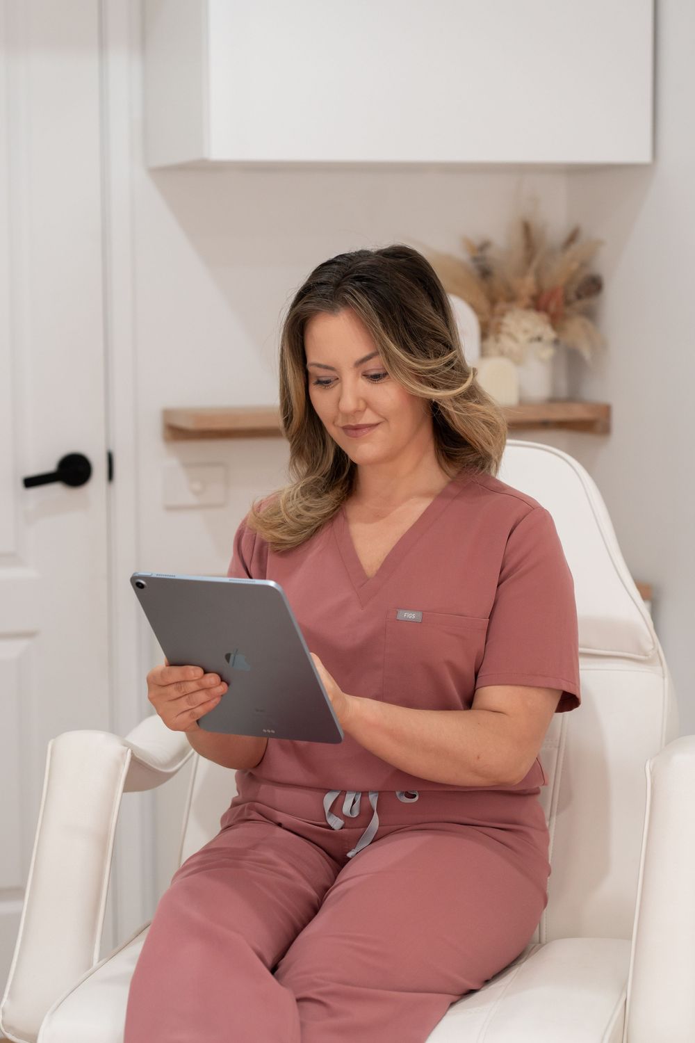 Healthcare professional in pink scrubs sitting in white chair reviewing patient information on a tablet device.