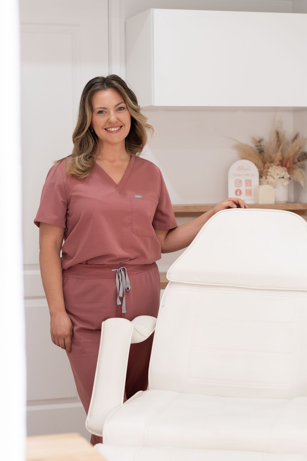 A smiling female healthcare professional in mauve scrubs stands beside a white treatment chair in a modern clinic.