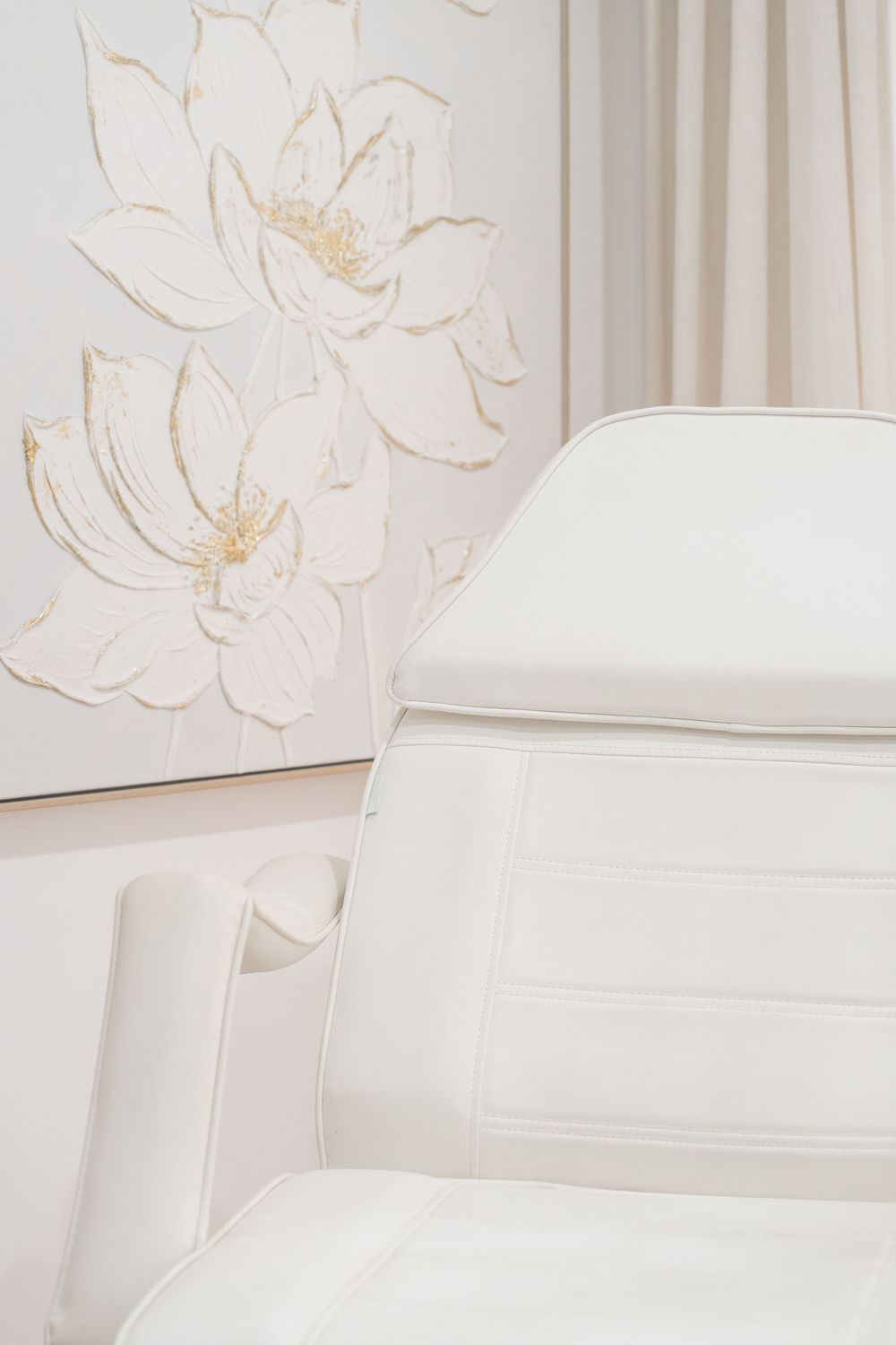 White leather spa treatment chair beside an elegant floral wall art with gold accents in a luxury beauty salon.