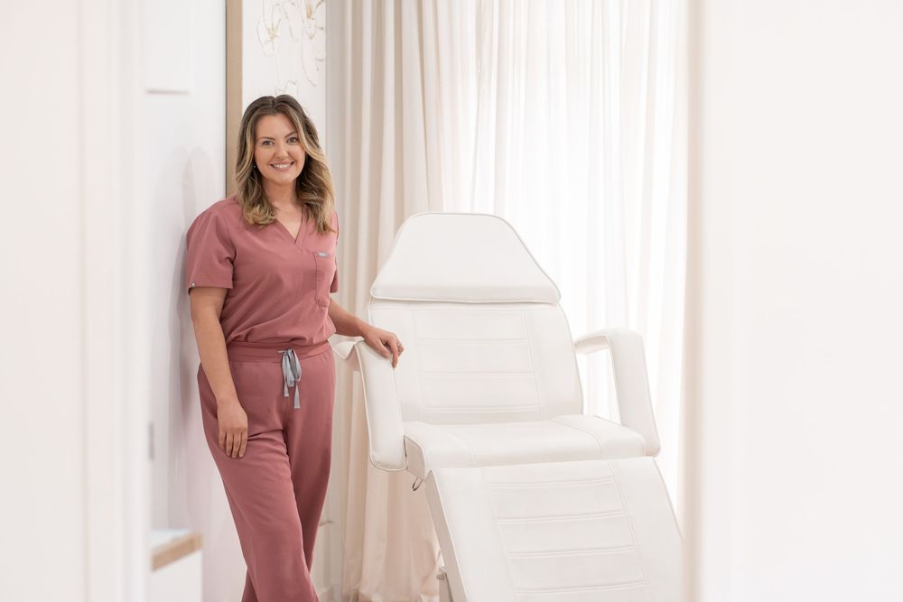 Woman in pink scrubs smiling beside a white treatment chair in a bright, elegant medical spa room.