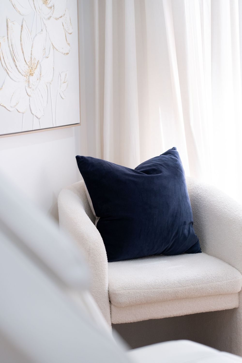White boucle armchair with navy blue velvet cushion beside sheer white curtains and floral wall art.