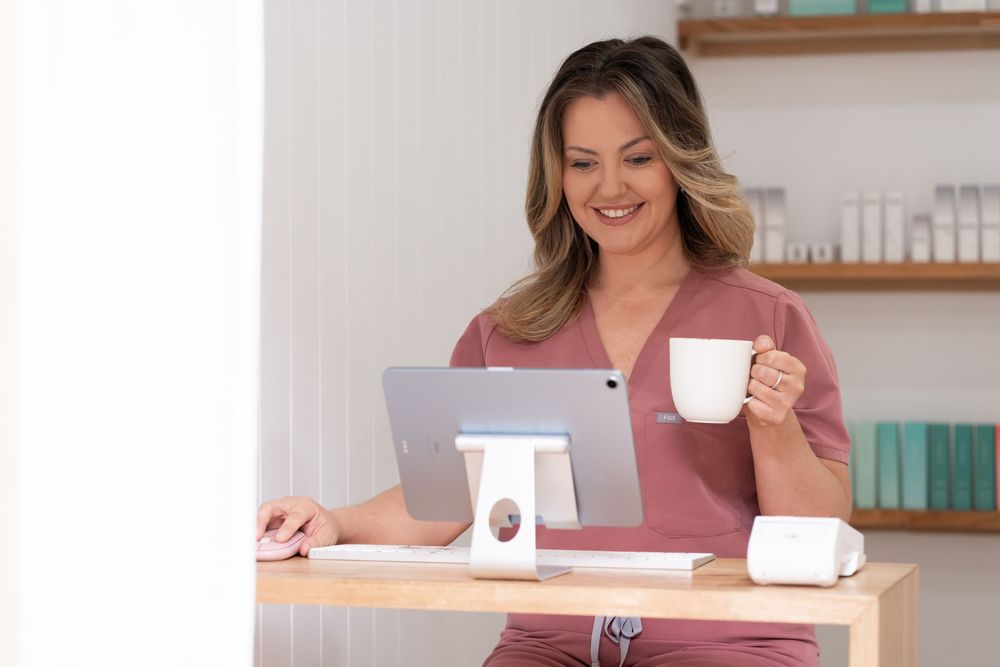 Woman in pink scrubs smiling at tablet while holding coffee cup at wooden desk in bright office setting.