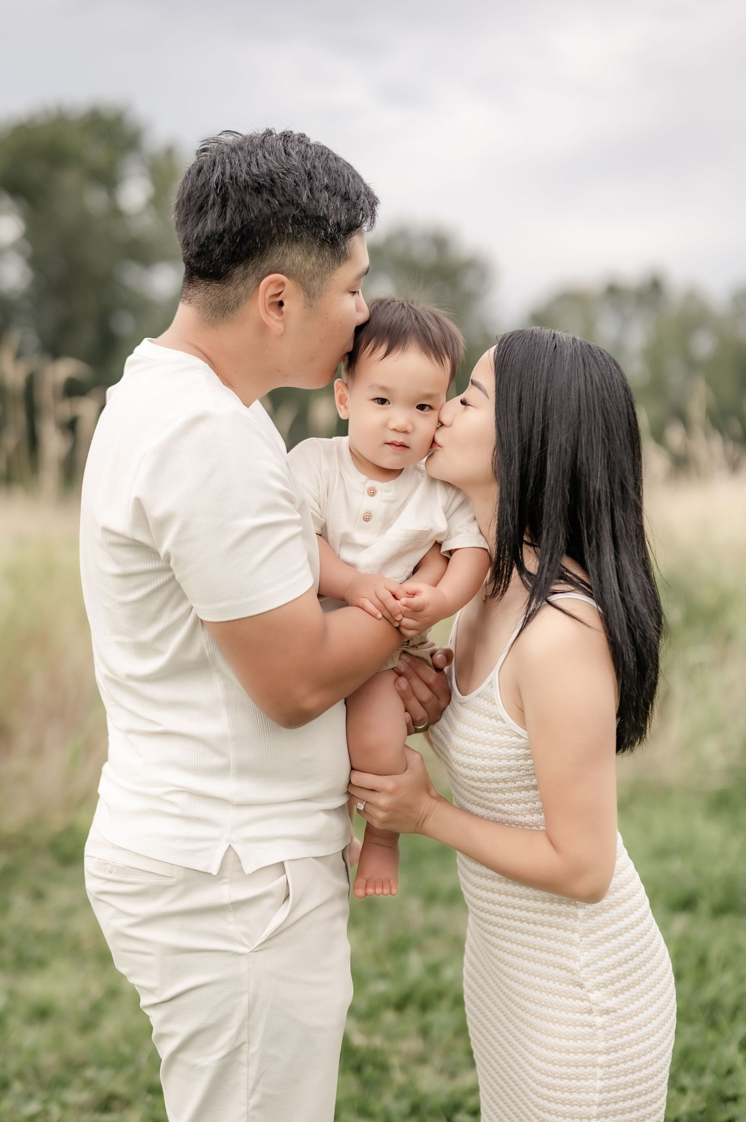 A couple dressed in white kisses their baby outdoors in a green field for a family portrait session.