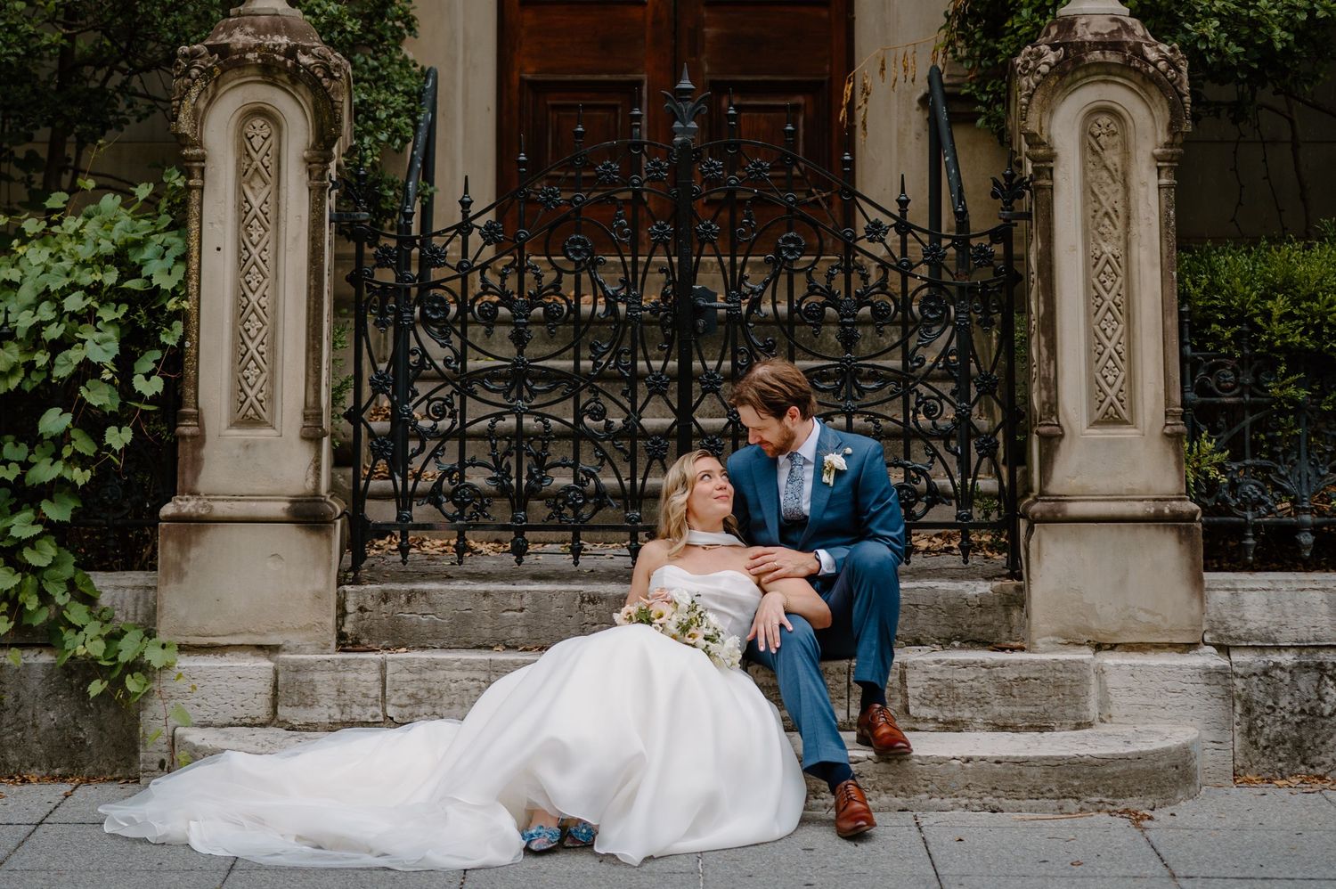 Bride and groom sitting on stone steps before ornate iron gate, bride in white gown, groom in blue suit.