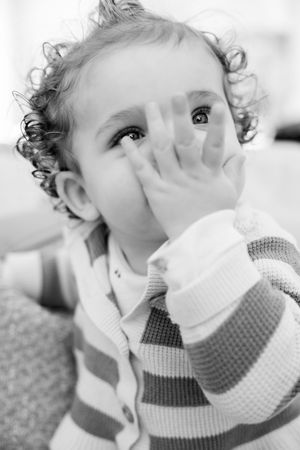 Black and white photo of a curly-haired toddler in a striped sweater covering mouth with hand, looking upward.