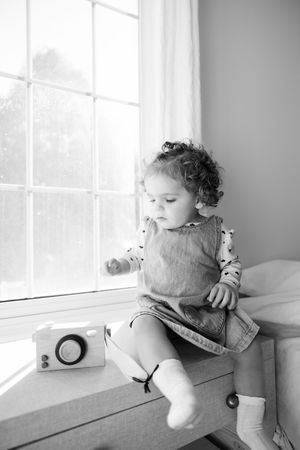 Black and white photo of a curly-haired toddler sitting on a window seat with a toy camera nearby.