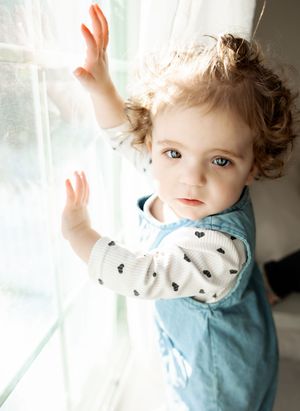 Curly-haired toddler with blue eyes reaching up toward a bright window, wearing a denim dress over a polka dot shirt.