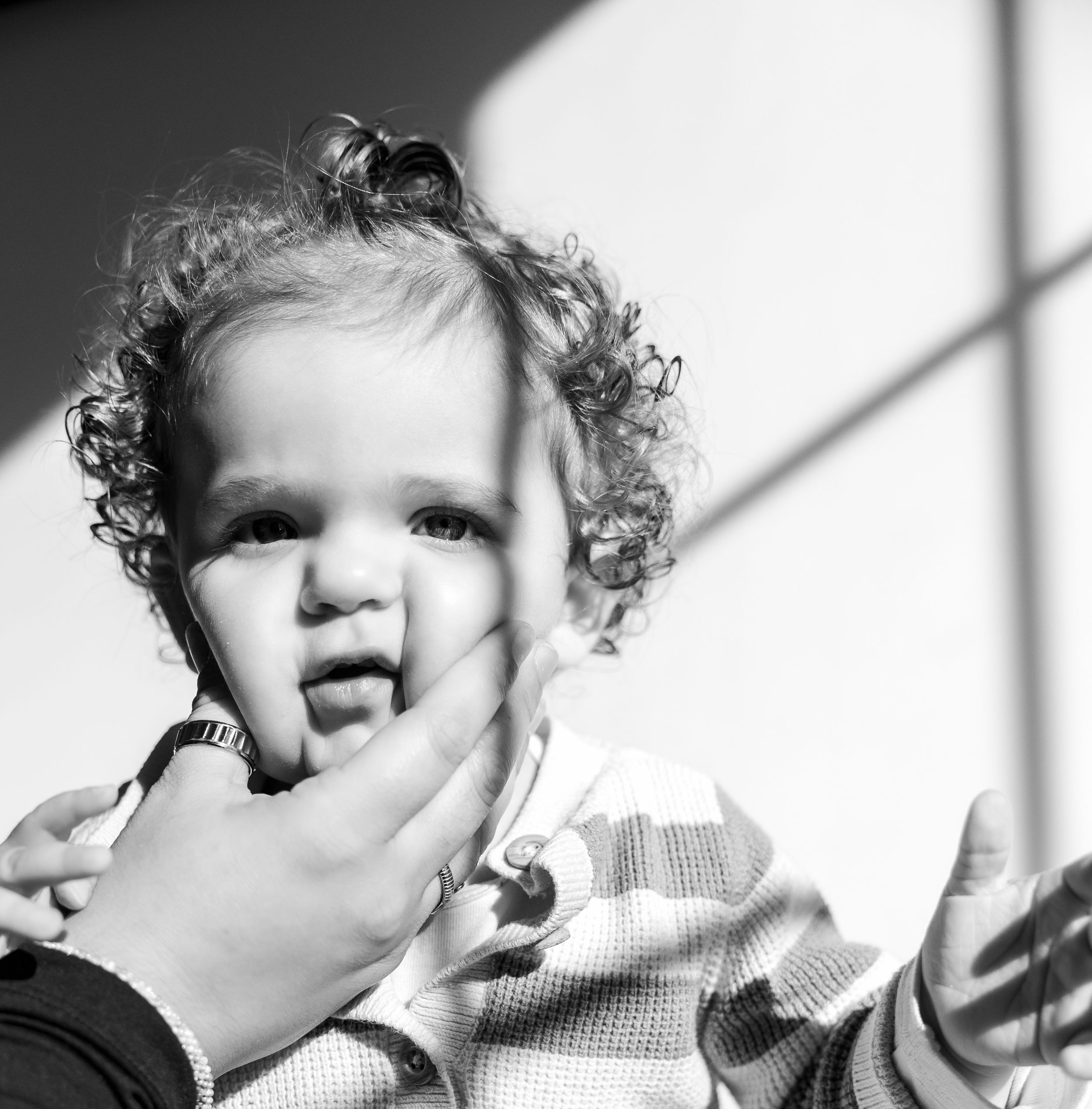 Black and white photo of a curly-haired toddler being held, looking curious with an adult hand gently touching their chin.
