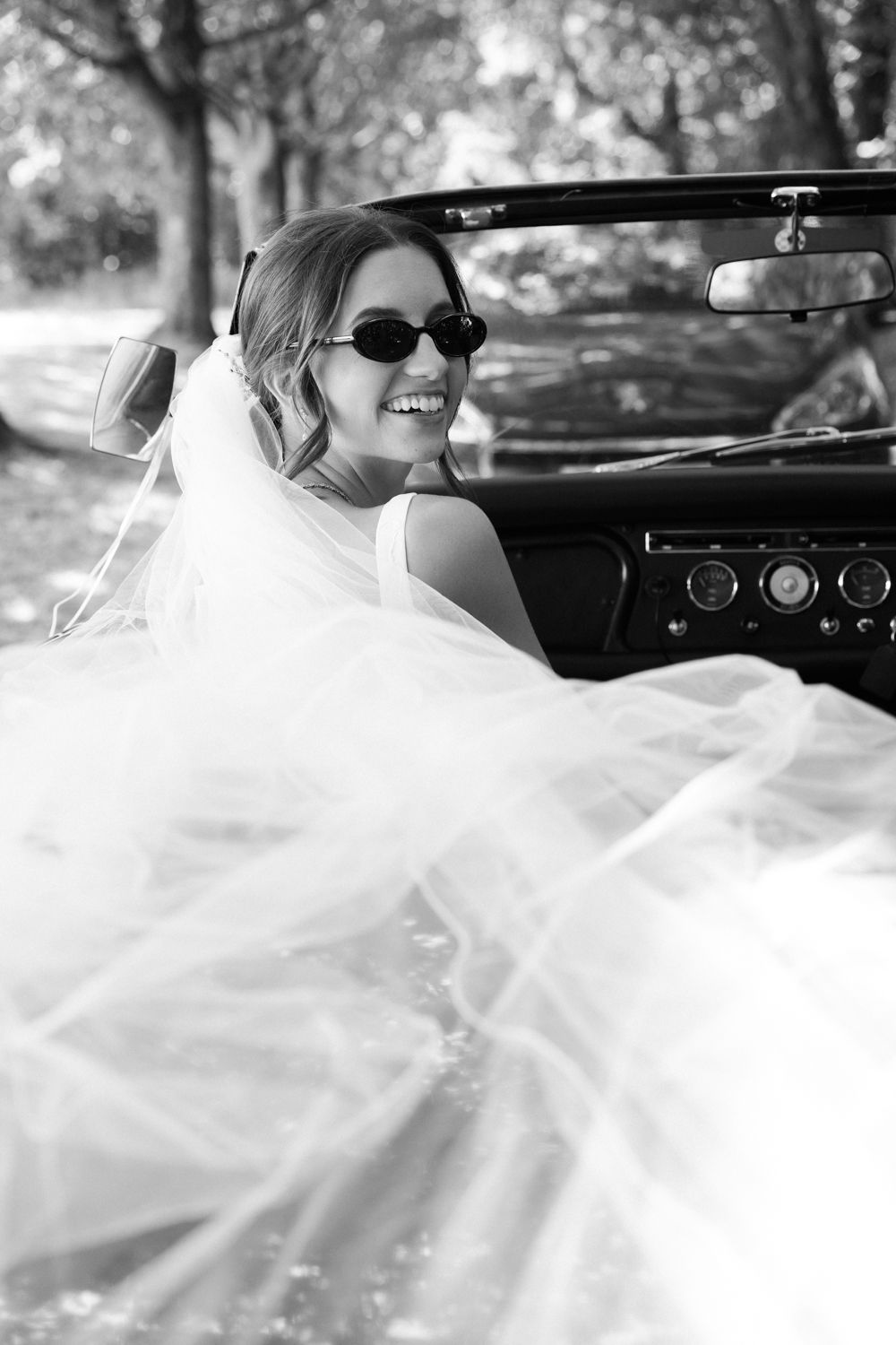 Smiling bride in sunglasses with flowing veil sits in vintage convertible car, black and white wedding photo.