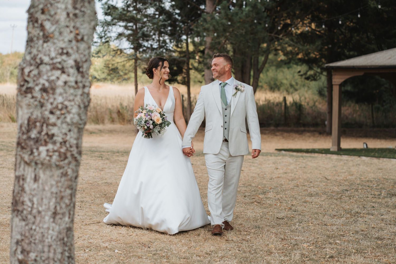 Bride and groom walking hand in hand outdoors on dry grass, bride holding bouquet in white gown, groom in light gray suit.