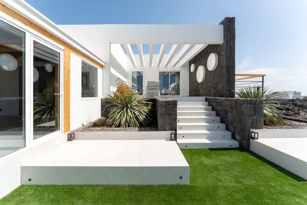 Modern white villa exterior with artificial grass, stone steps, pergola, and tropical plants under a clear blue sky.