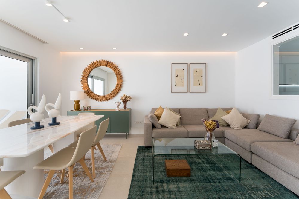 Modern open-plan living room with beige sofa, green rug, white dining set, and sunburst mirror on white walls.