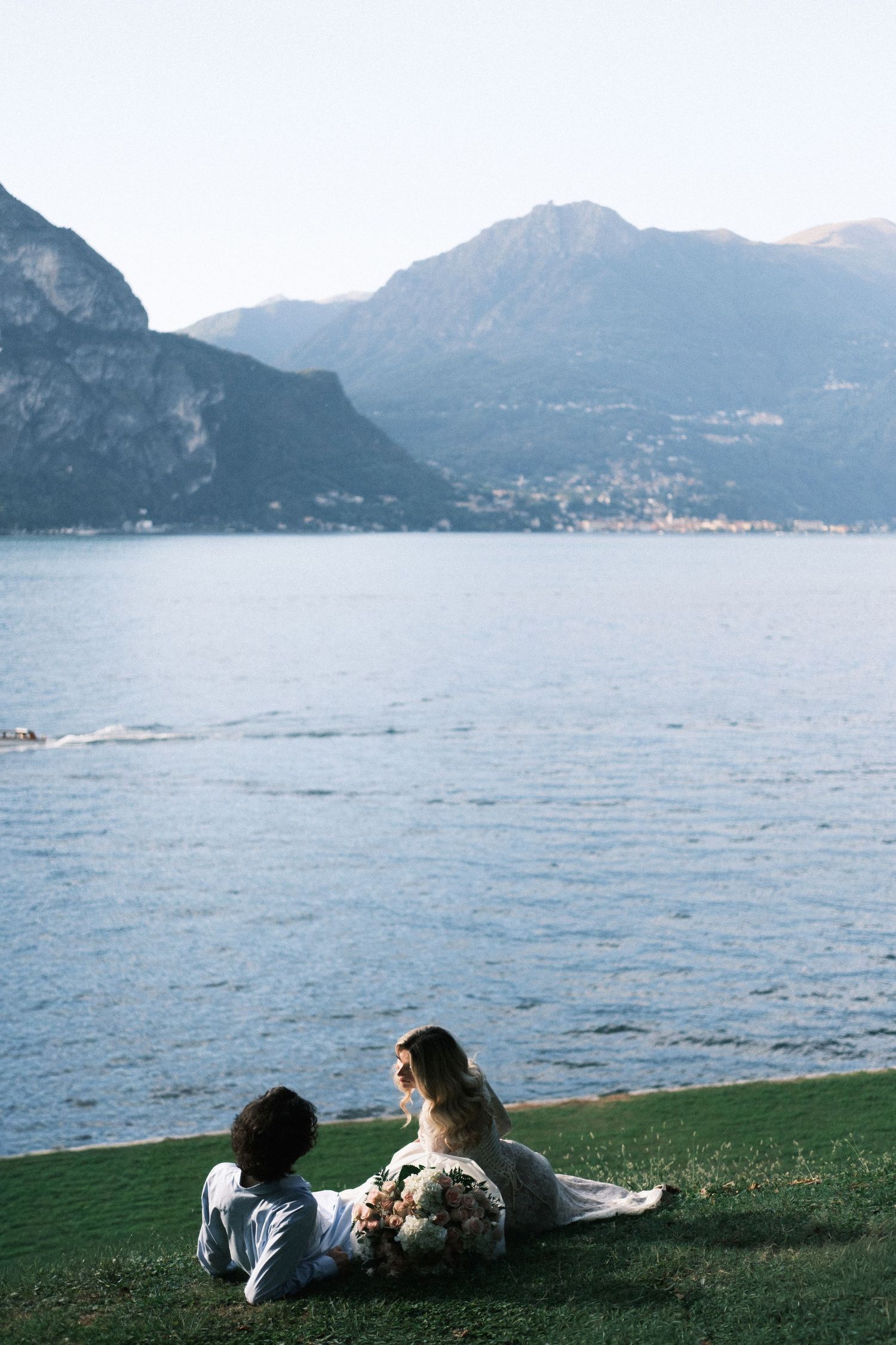 Bride and groom sitting on green grass by a serene lake with mountain backdrop on their wedding day.
