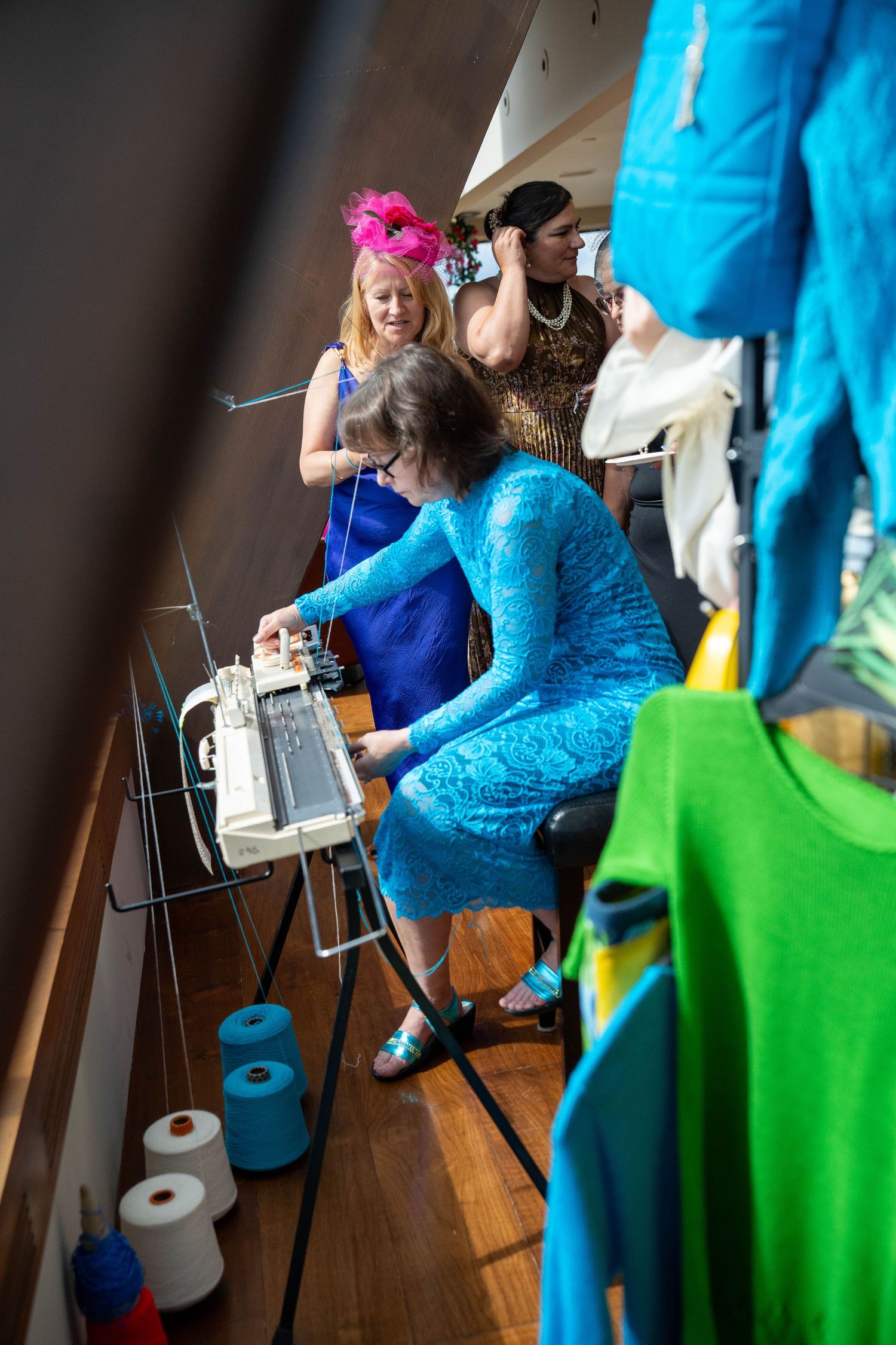 Women in colorful dresses gather around a knitting machine with yarn spools on a wooden floor.