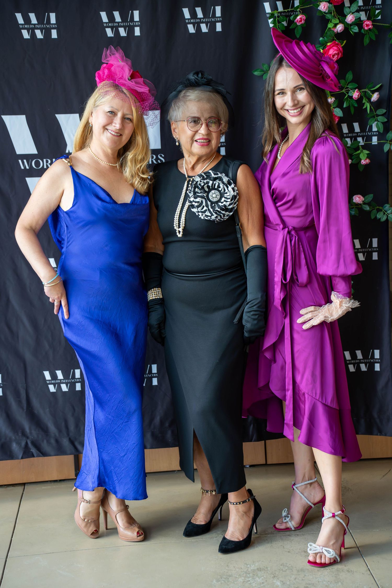 Three elegantly dressed women in colorful gowns and decorative hats pose together at a formal event.