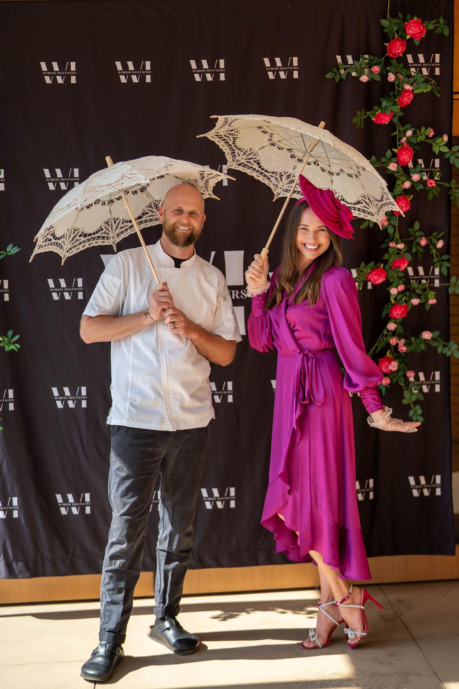 A man and woman pose with lace parasols in front of a floral backdrop, she wearing a magenta dress and hat.