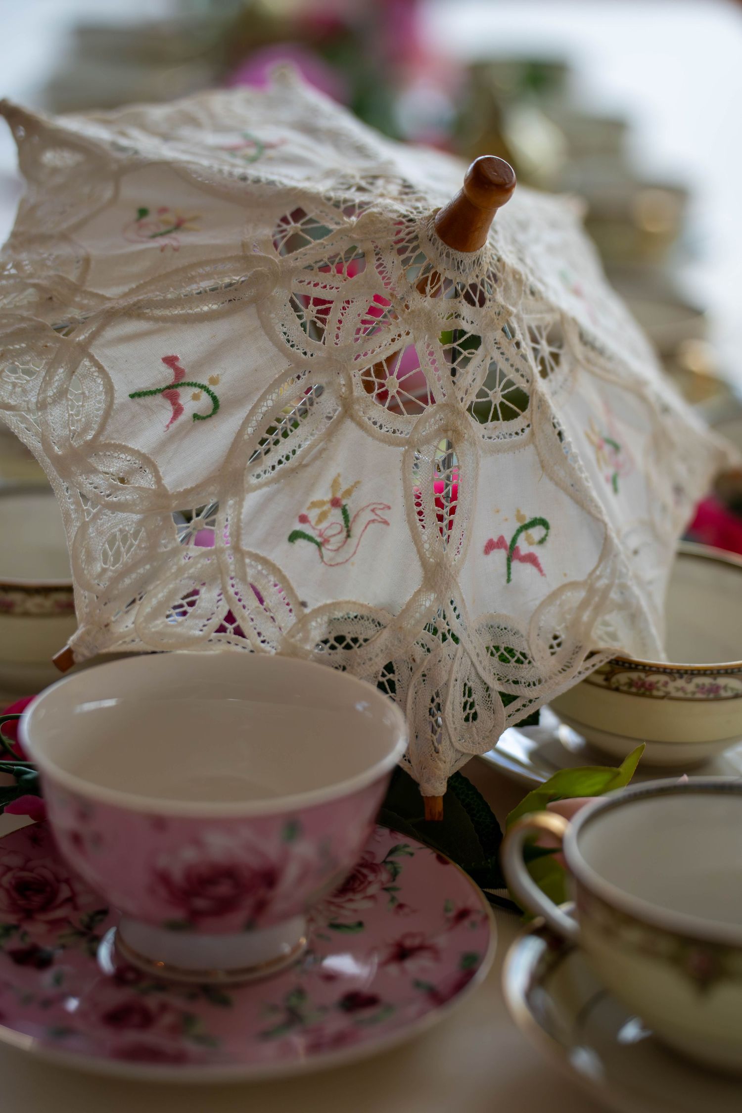 Vintage lace parasol surrounded by floral china teacups and saucers arranged on a table for an elegant tea party.