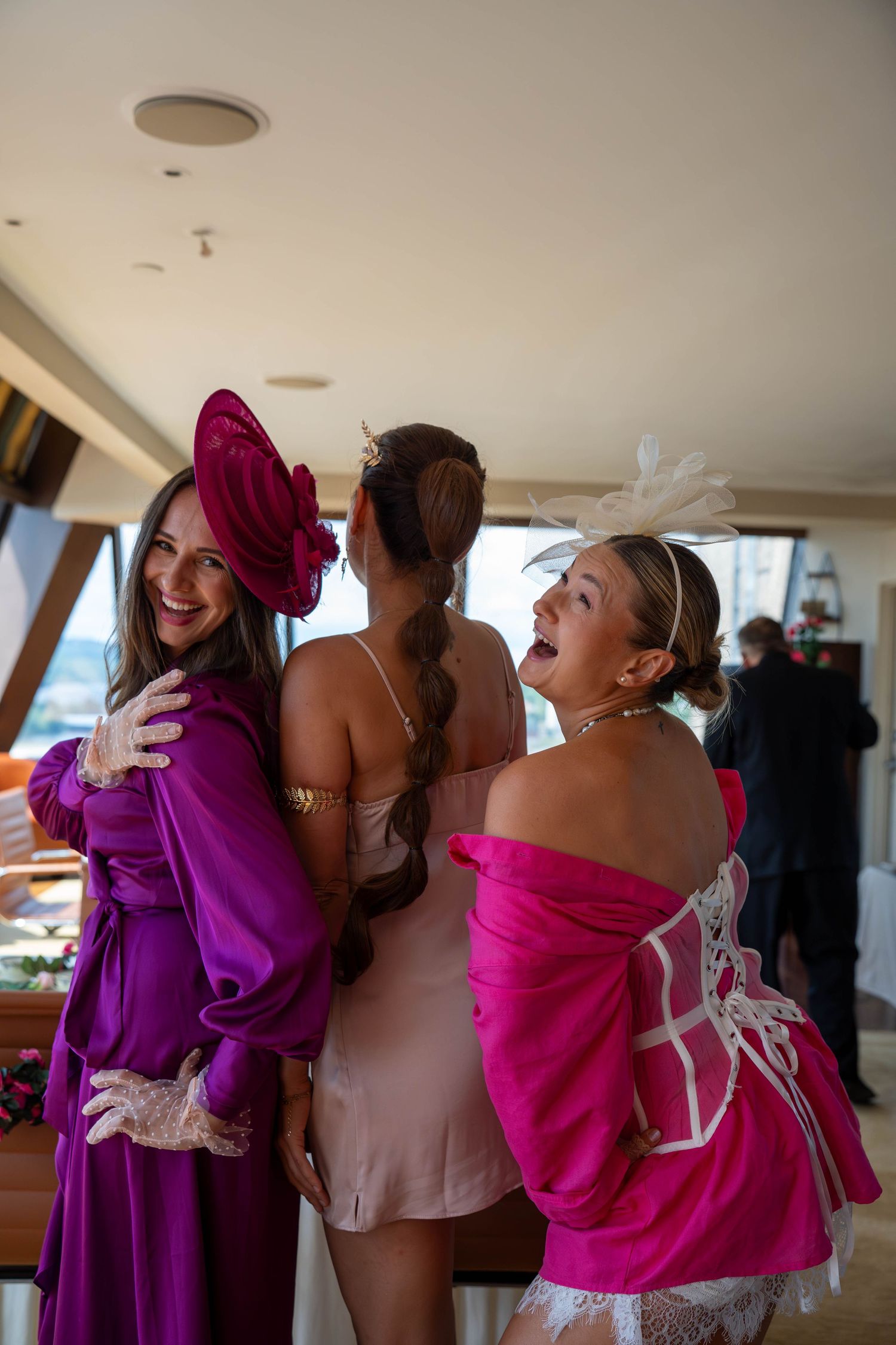 Three women in vibrant pink and purple formal outfits with fascinators laughing together at an elegant event.
