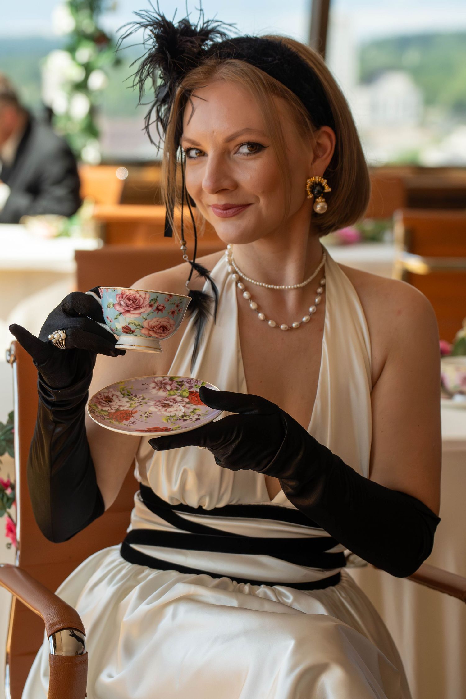 Elegant woman in white gown, black gloves, pearl necklace, and fascinator sipping tea from floral cup and saucer.