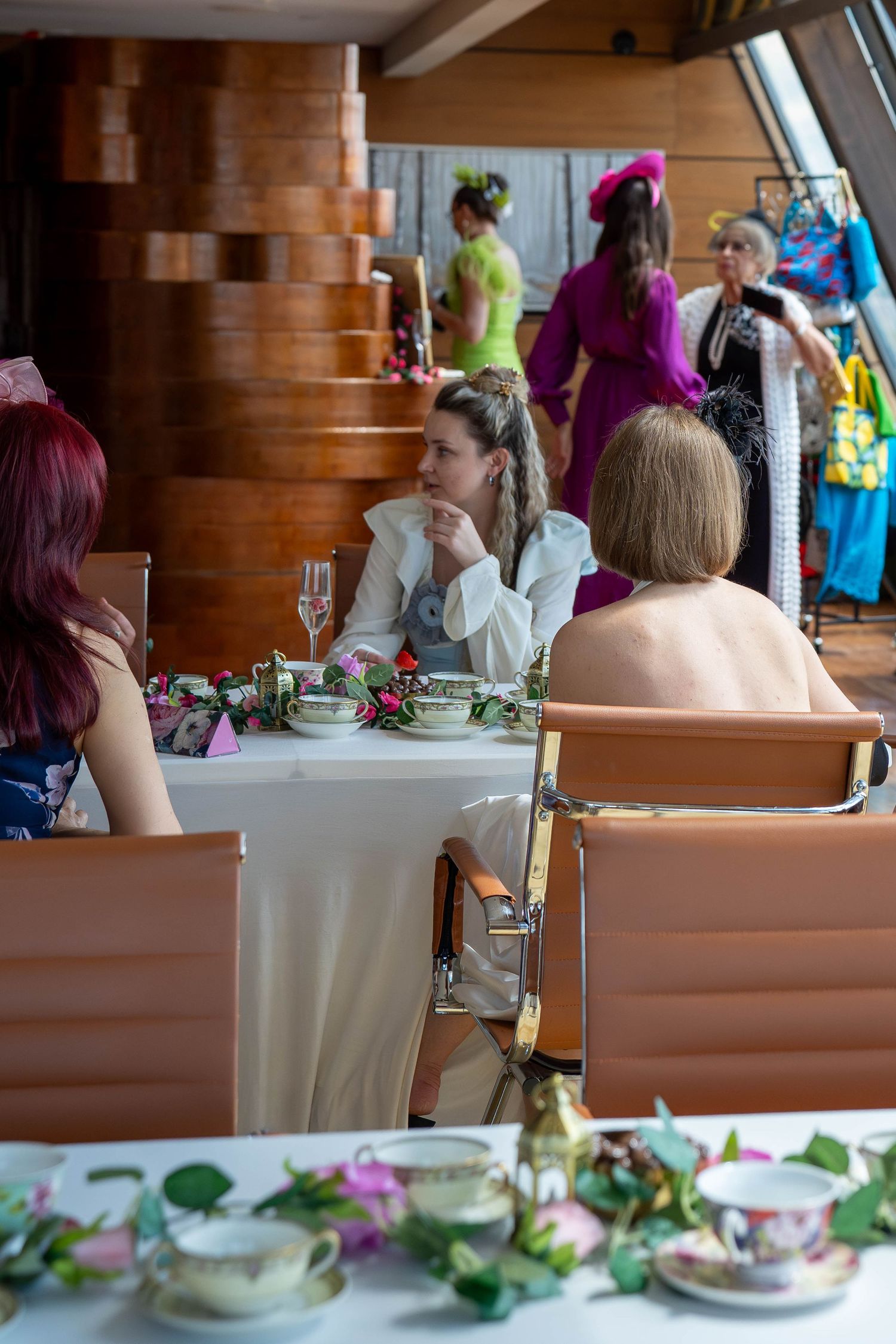 Women seated at a floral-decorated table during an elegant fashion event with colorfully dressed models in the background.