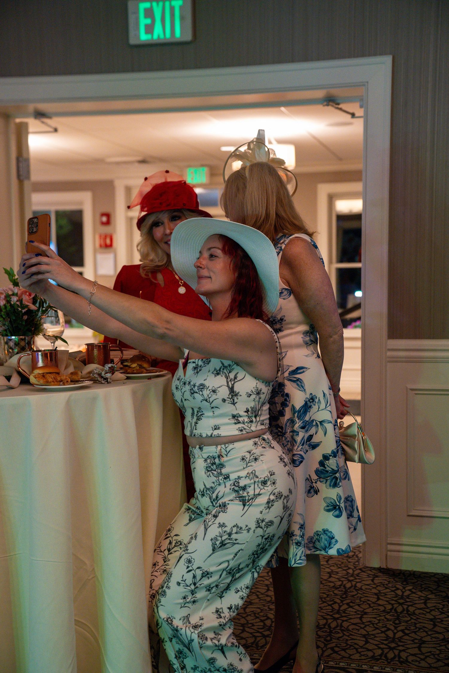 Three women in elegant dresses and decorative hats take a selfie at an indoor event near a buffet table.