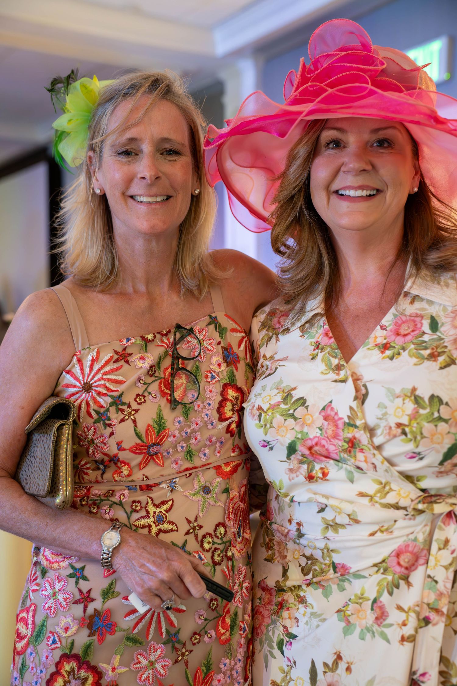 Two smiling women in floral dresses and decorative hats pose together at a festive garden party event.
