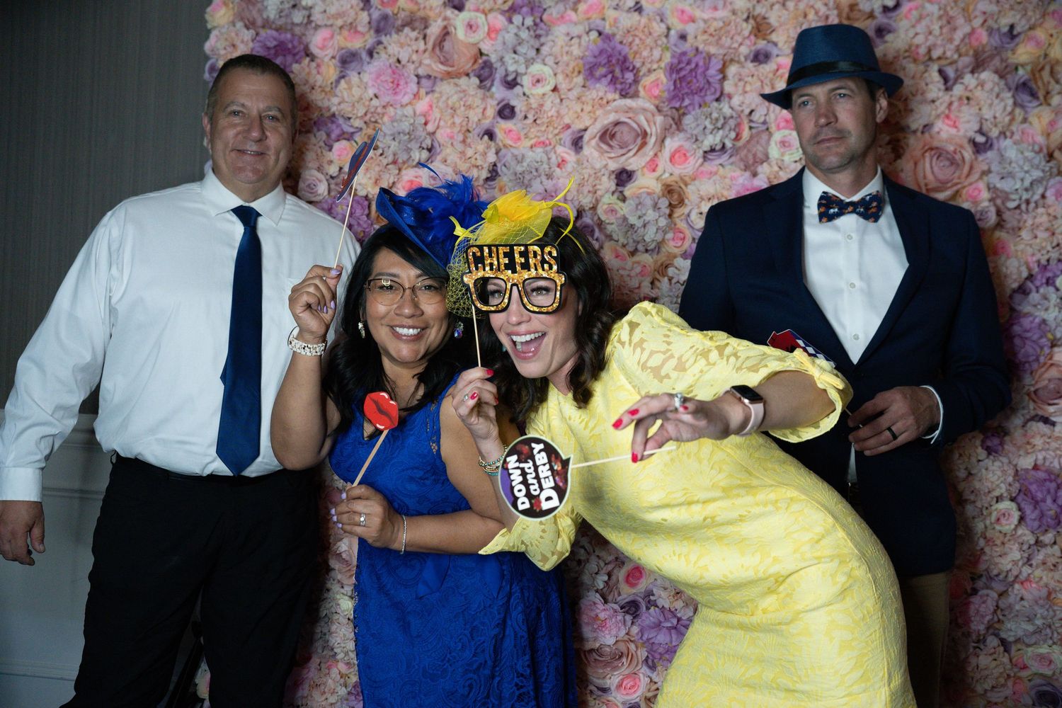 Four friends pose with fun props in front of a floral wall backdrop at a festive event.