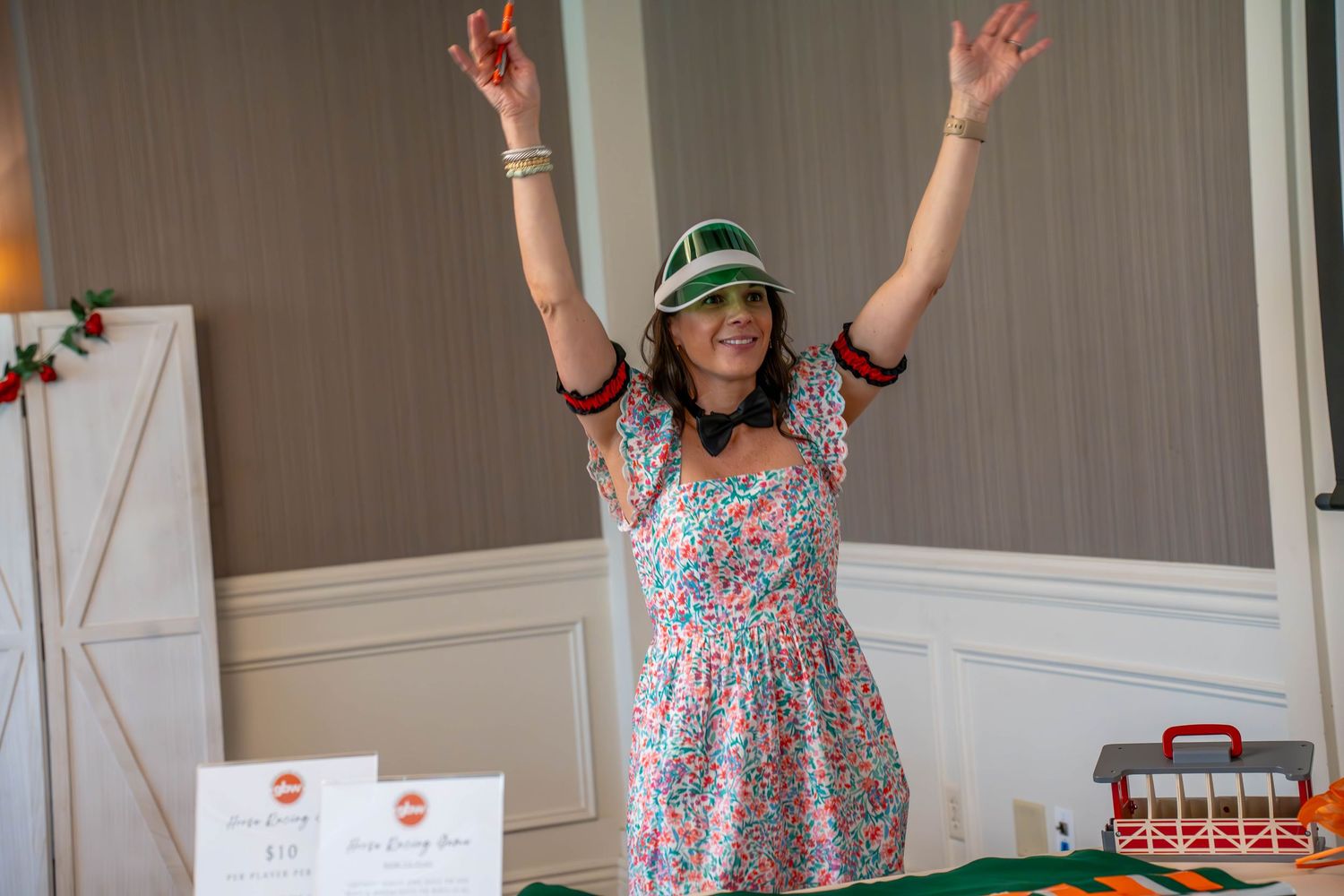 Woman in floral dress and green visor raises arms in celebration at a festive holiday event table.