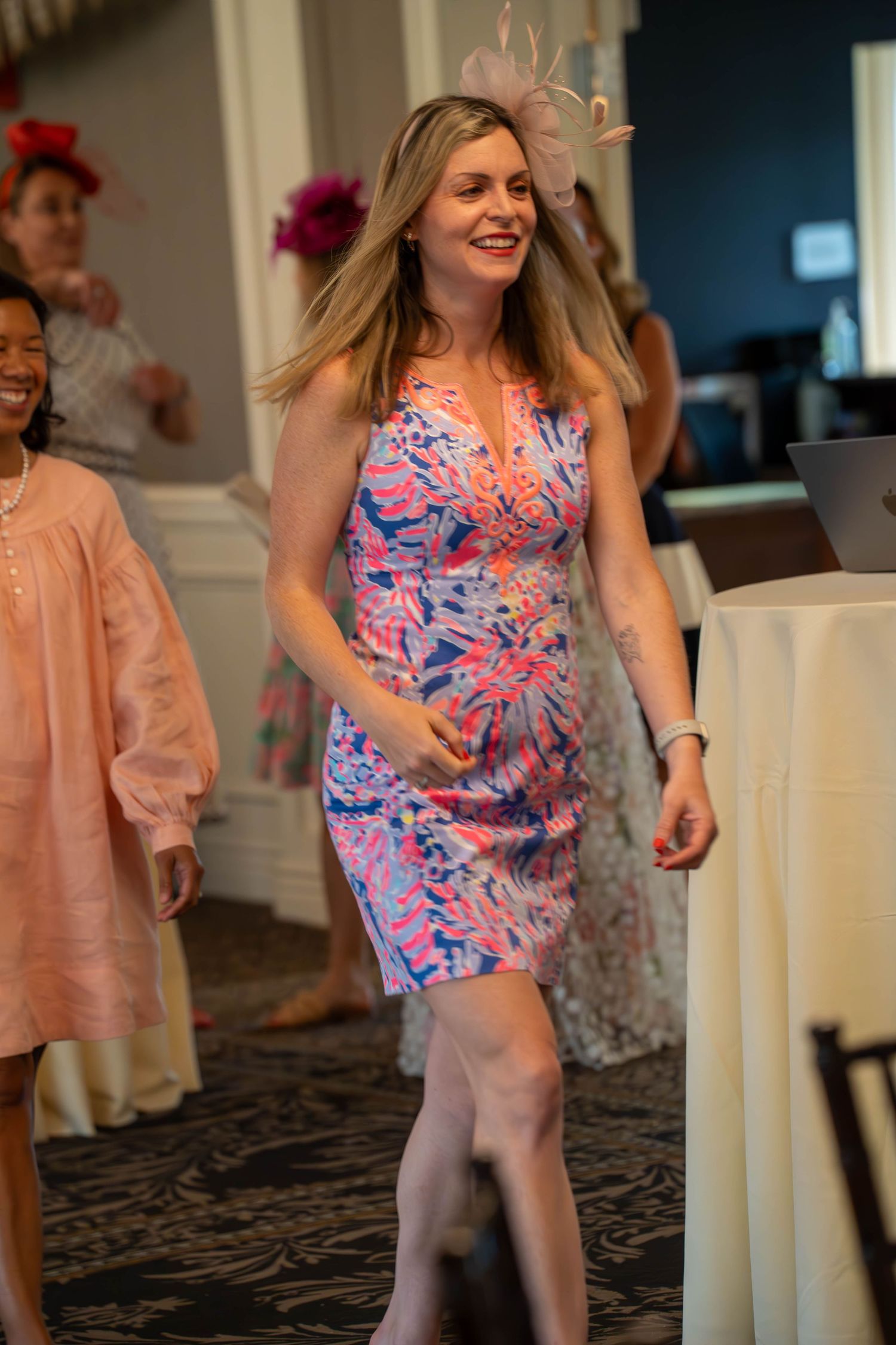 Woman in floral pink dress and fascinator hat walking at an elegant indoor event or party.