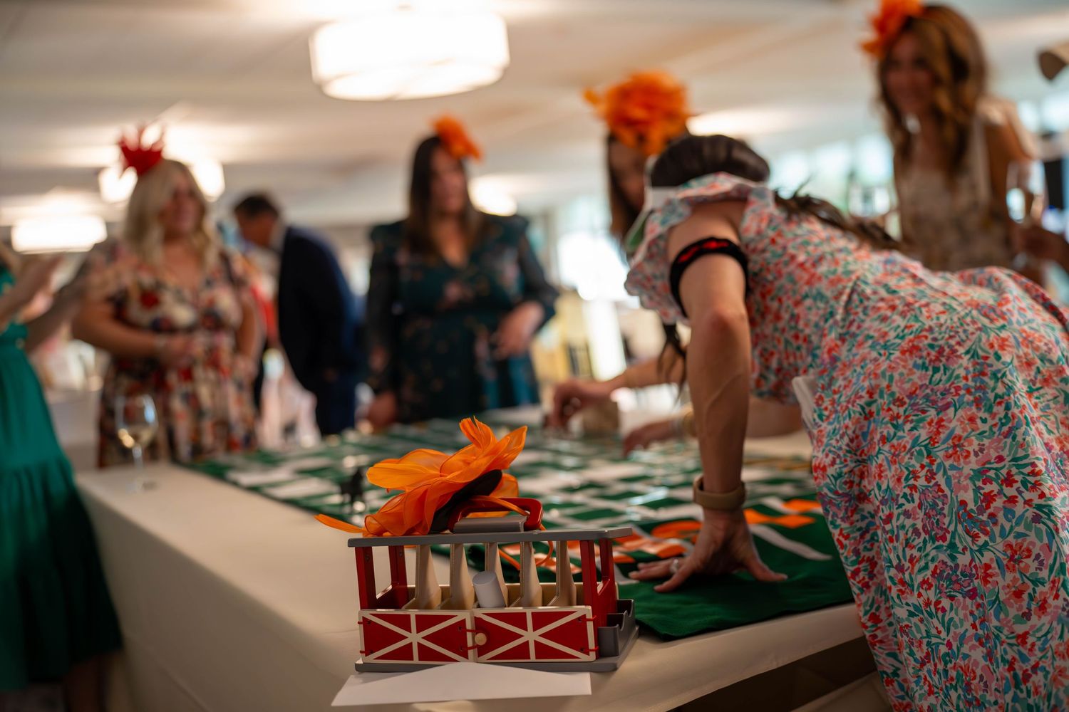 Women in floral dresses and orange fascinators play a toy horse racing game at an elegant indoor event.