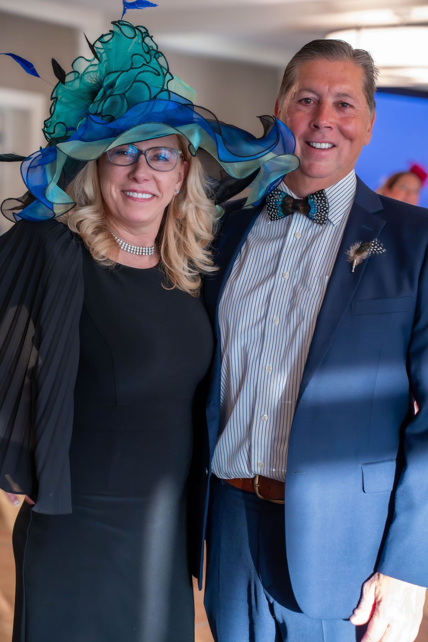 Elegantly dressed couple posing at formal event, woman in teal fascinator hat and black dress, man in navy suit with bow tie.