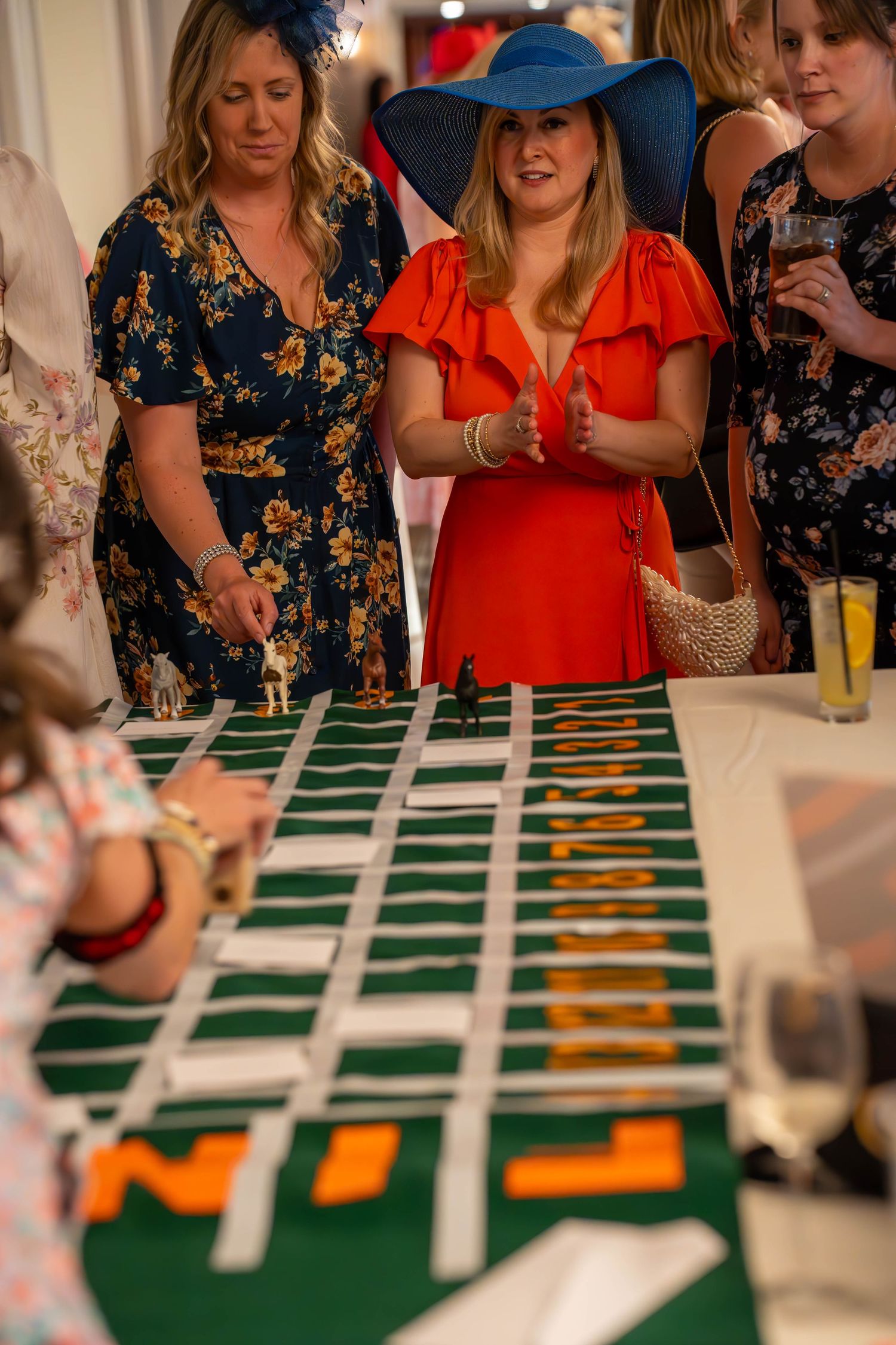 Women in colorful dresses and wide-brim hats play a tabletop horse racing game at a Kentucky Derby party.