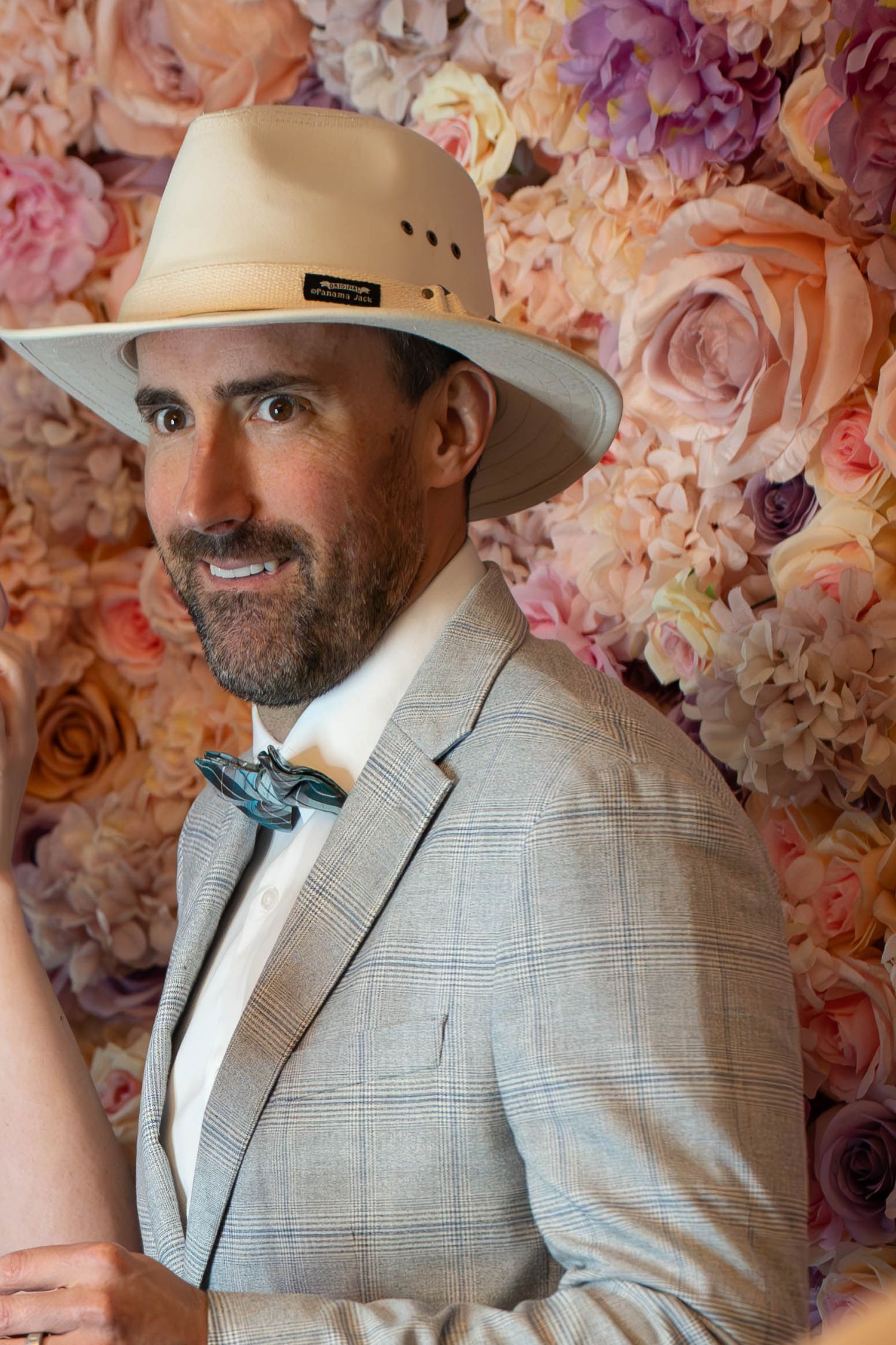Man in tan wide-brim hat and plaid suit with teal bow tie posing before a floral wall backdrop.