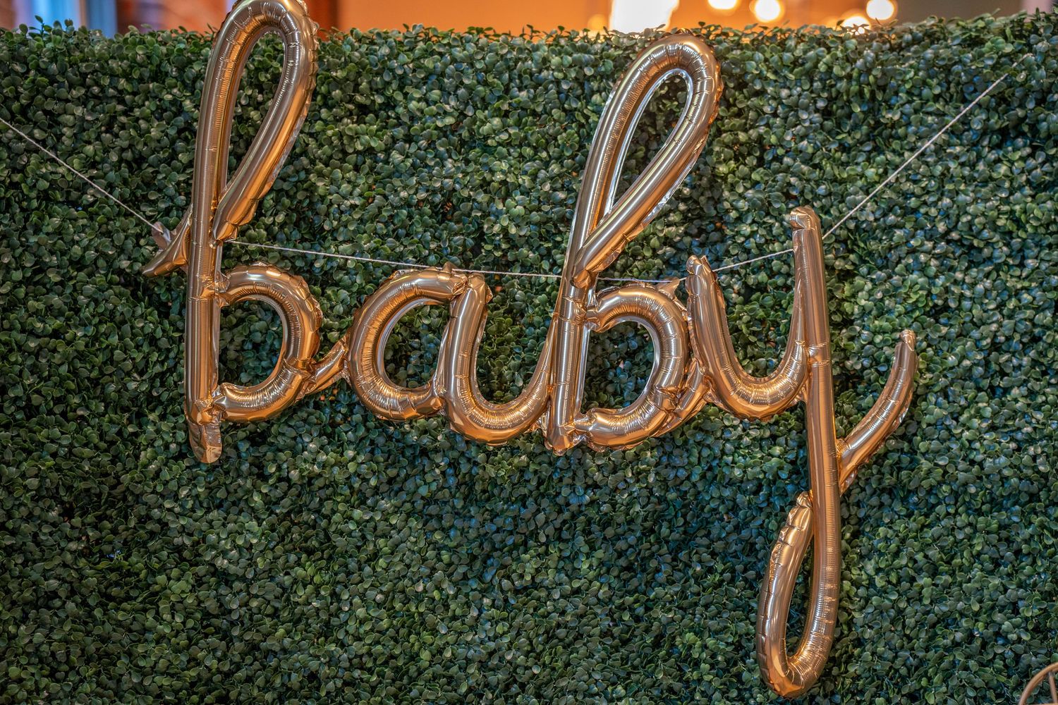 Gold script balloon letters spelling baby against a lush green boxwood hedge wall at a baby shower.