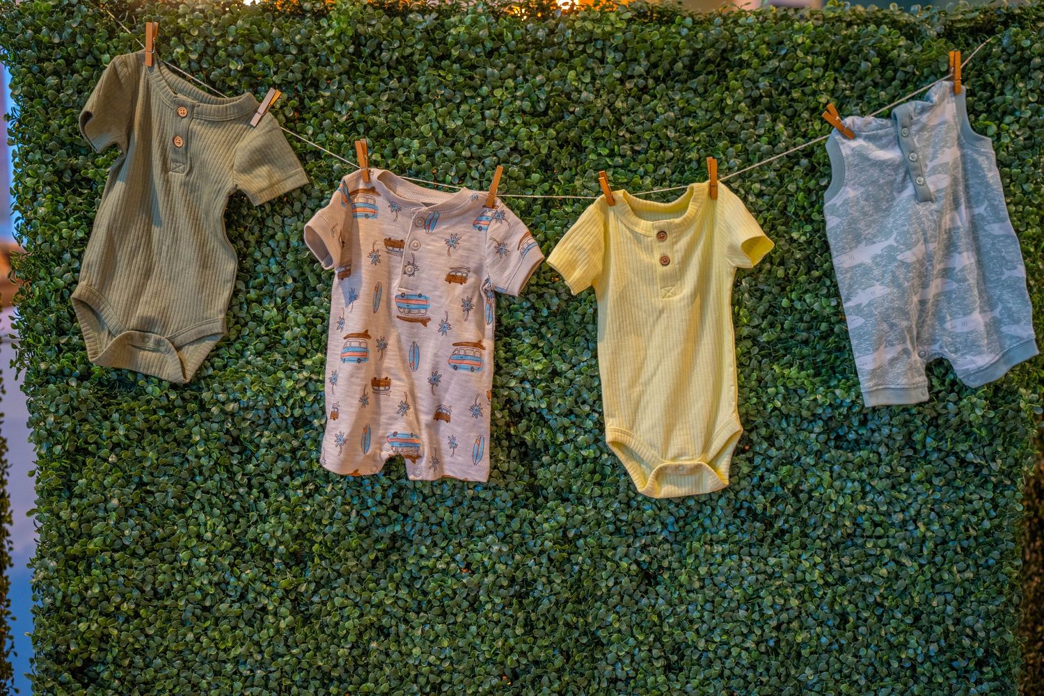 Four colorful baby onesies hanging on a clothesline against a lush green ivy wall backdrop.