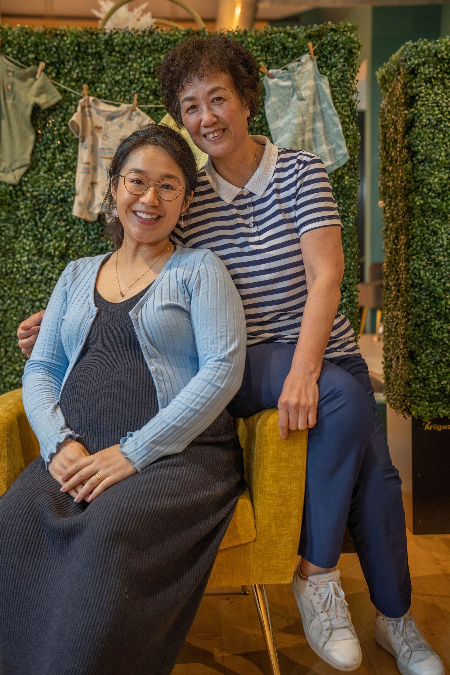 Two smiling Asian women pose together in a cozy shop with green wall backdrop, one visibly pregnant in blue cardigan.