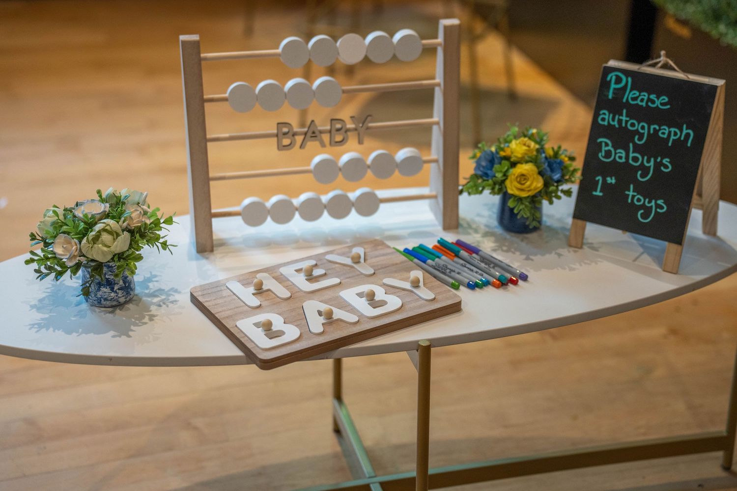 Baby shower gift table with wooden abacus, toy blocks spelling BABY, colorful markers, floral arrangements, and chalkboard sign.