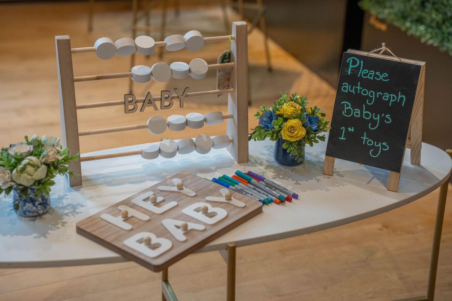 Baby shower guest sign-in table with wooden abacus, HEY BABY sign, markers, and chalkboard asking guests to autograph baby's first toys.